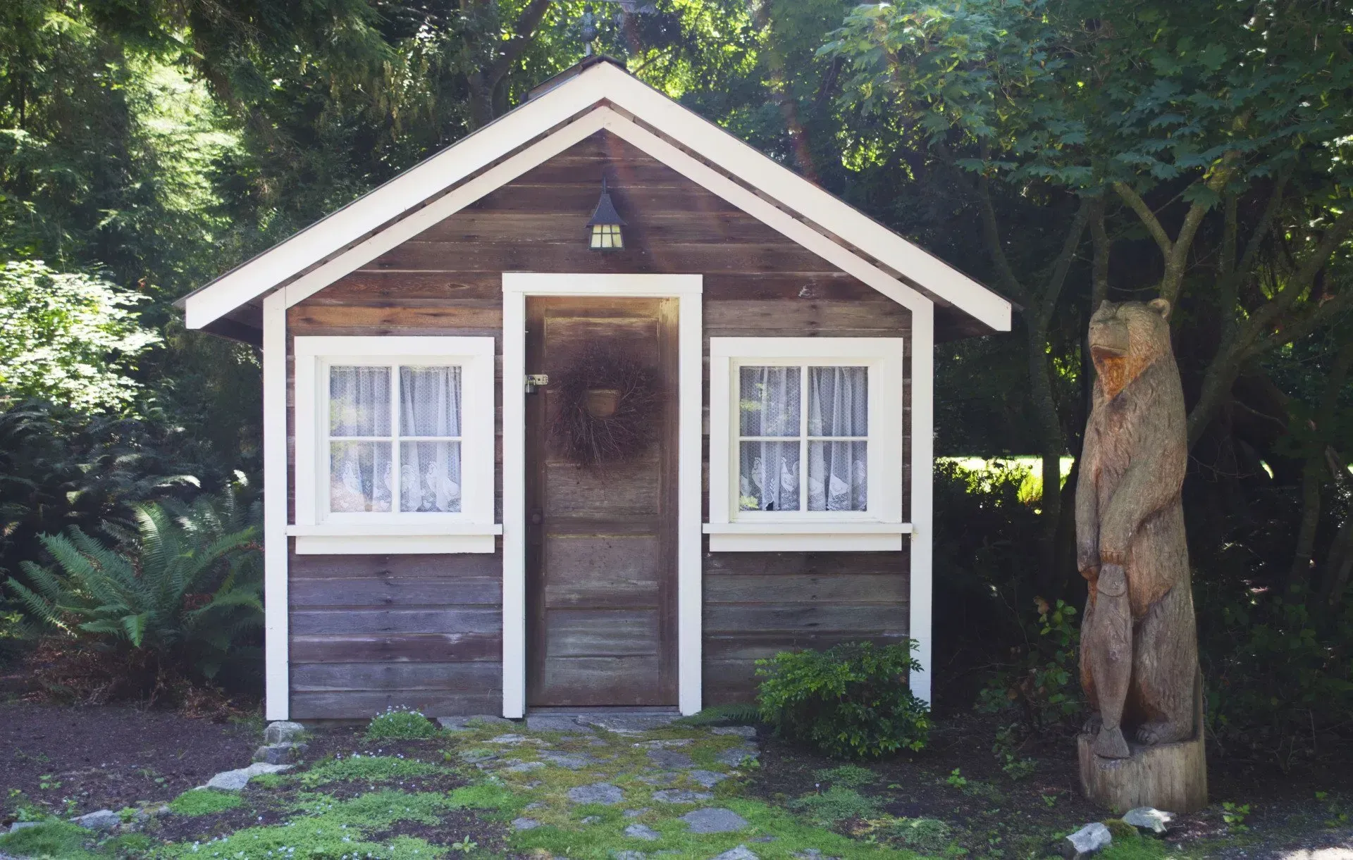 Small wooden shed with a white-trimmed door and windows, a carved bear statue, and foliage.