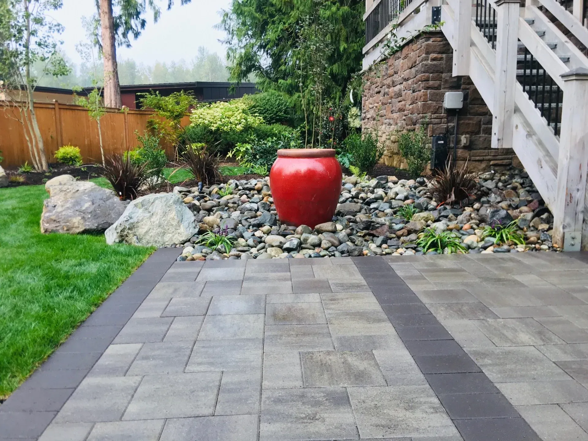 Patio with brick pavers, grass, red pot on rocks, and a wooden deck.