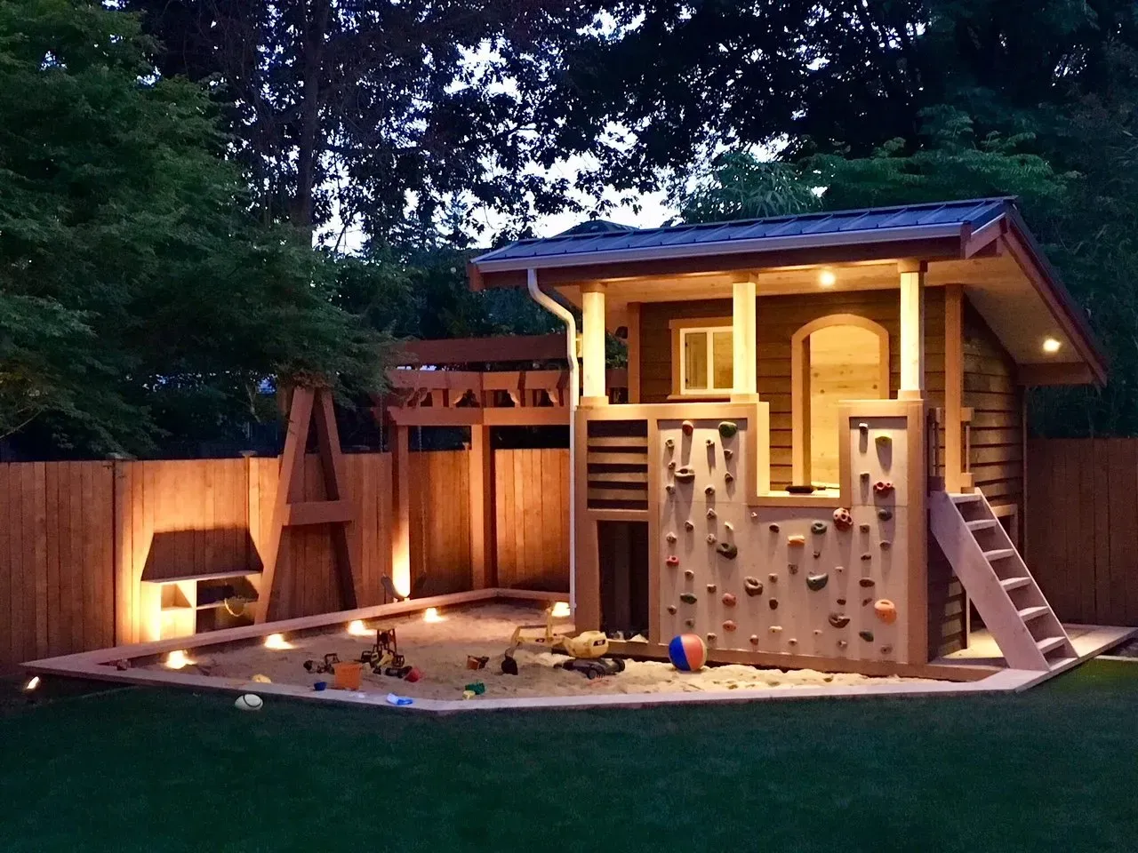 Playhouse with climbing wall, sandbox, and wooden fence illuminated at dusk.