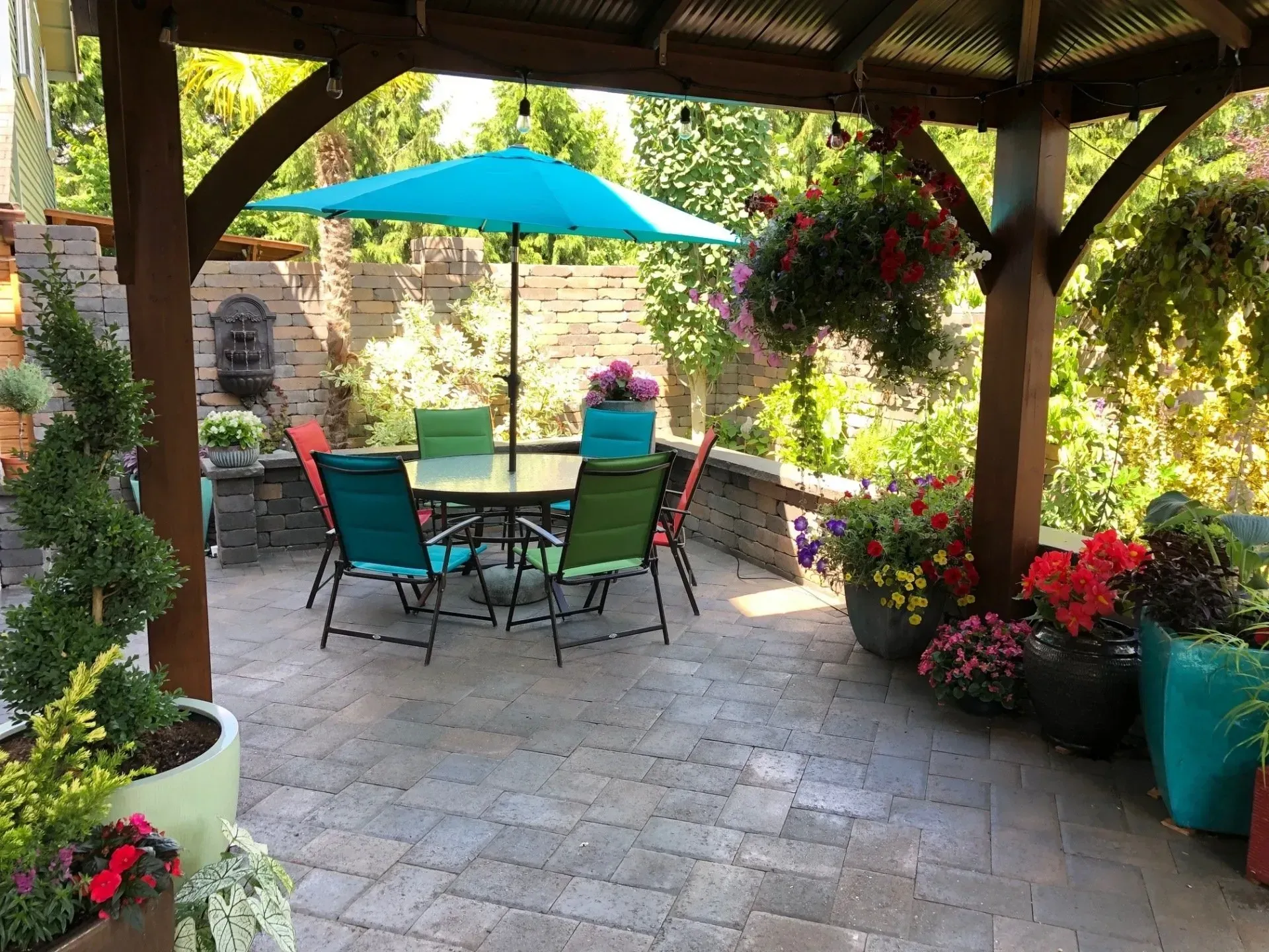 Patio with a table and chairs under an umbrella, surrounded by plants and a gazebo.