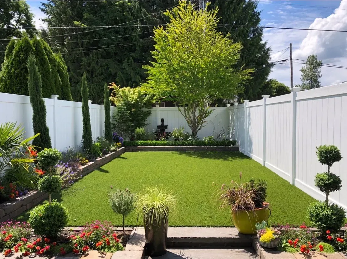 Lush green backyard with artificial turf, bordered by white fence, colorful flowers, and manicured shrubs. Sunny day.