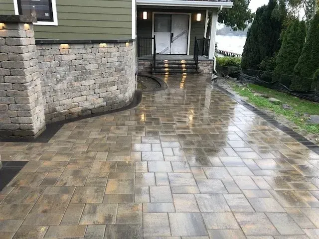 Paver driveway leading to a house entrance, bordered by a stone retaining wall and green shrubs.