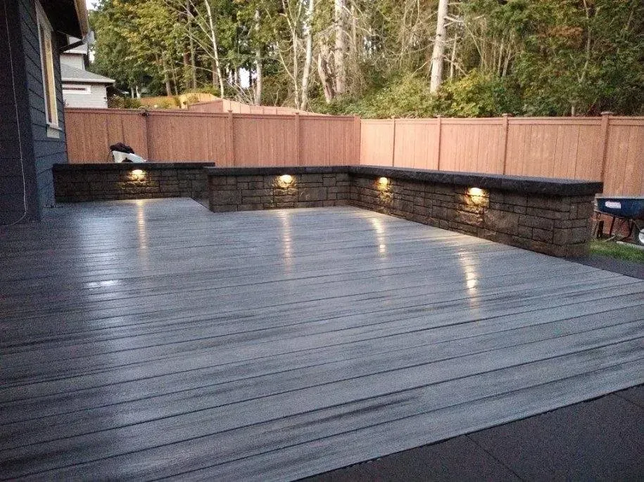 Outdoor deck with stacked stone retaining walls, lit by spotlights, backed by a wooden fence and trees.