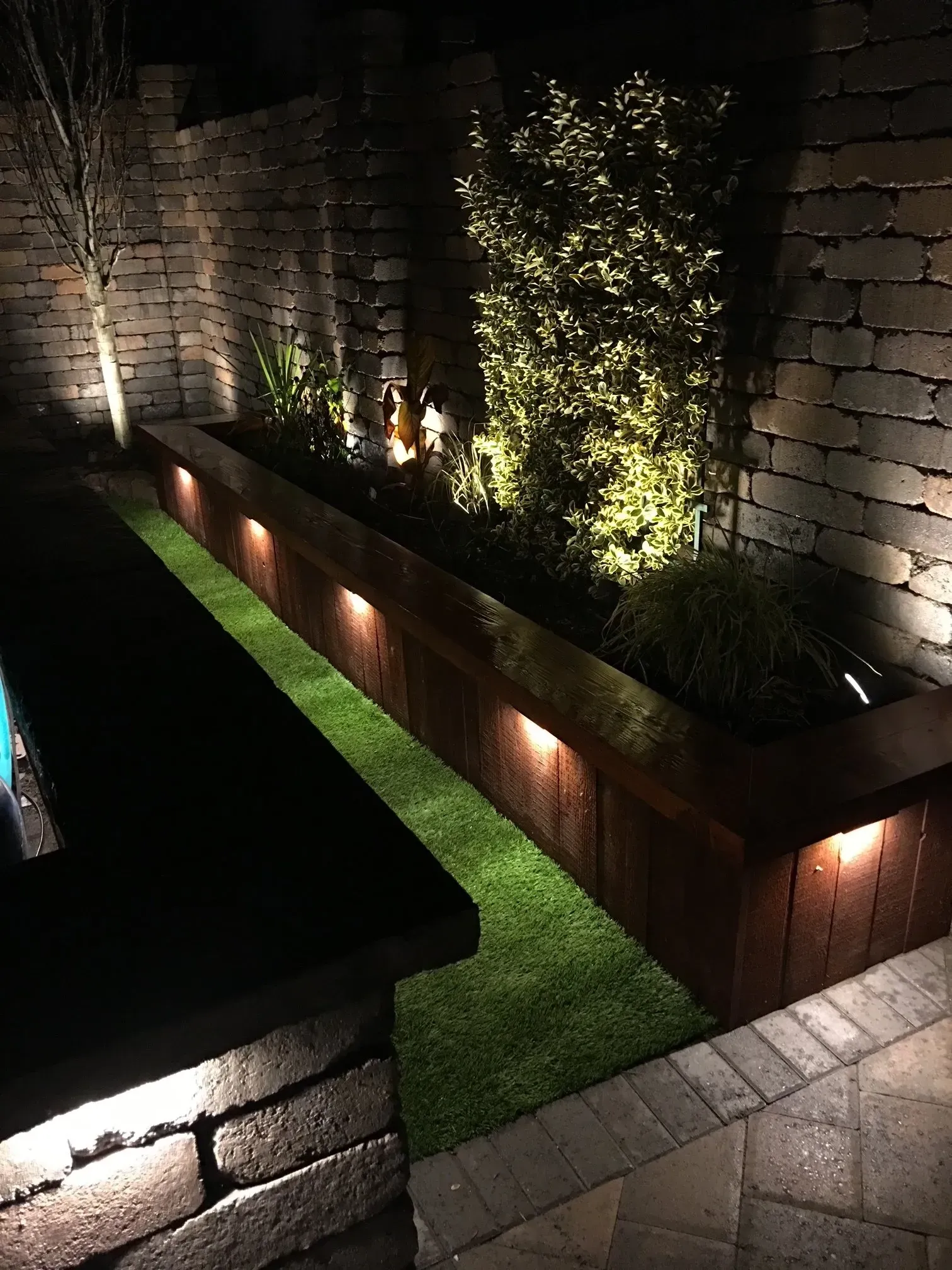 Nighttime view of a long wooden planter box with built-in lights, filled with green plants, on artificial turf.