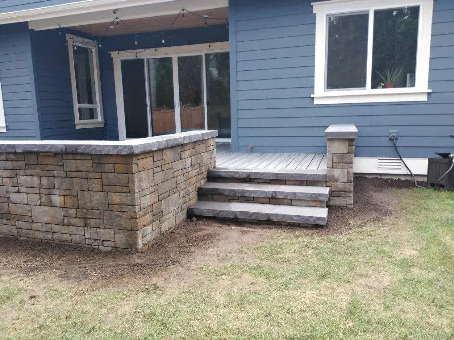 Stone patio with steps and retaining wall in front of a blue house with a sliding glass door and window.