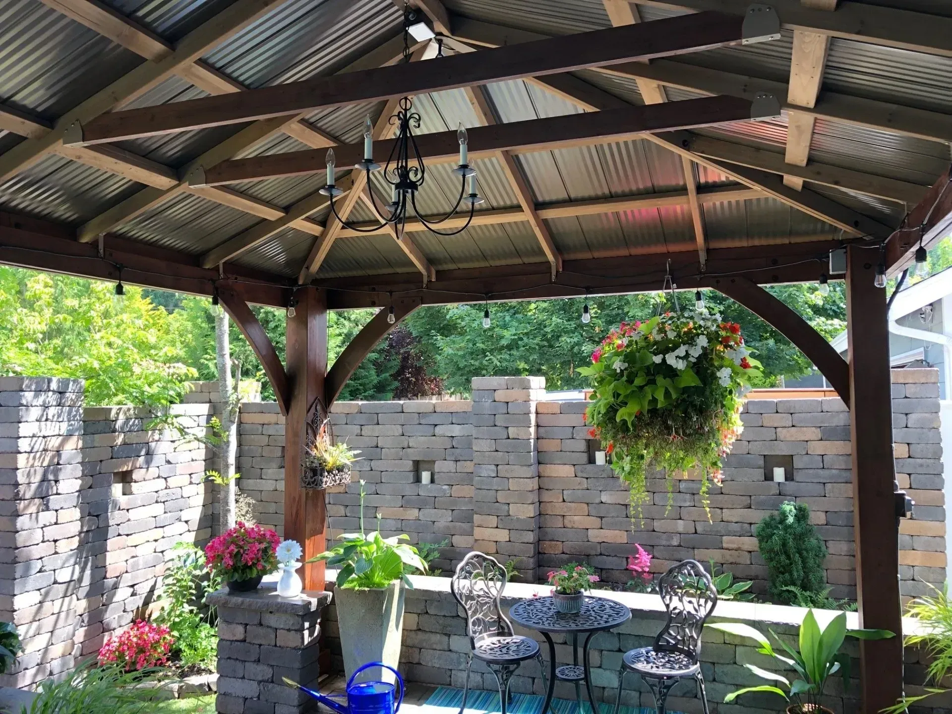 Gazebo with metal roof, wooden beams, chandelier, flowers, and a table with chairs.