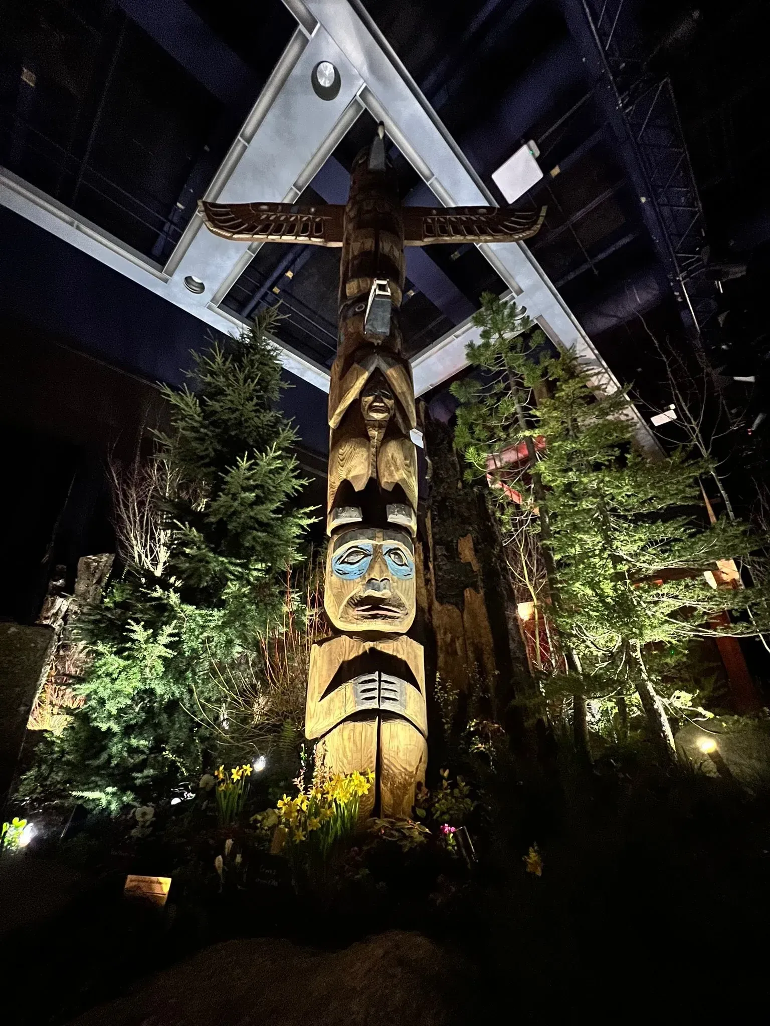 Totem pole in a dimly lit, indoor exhibit with surrounding evergreen trees and dark background.