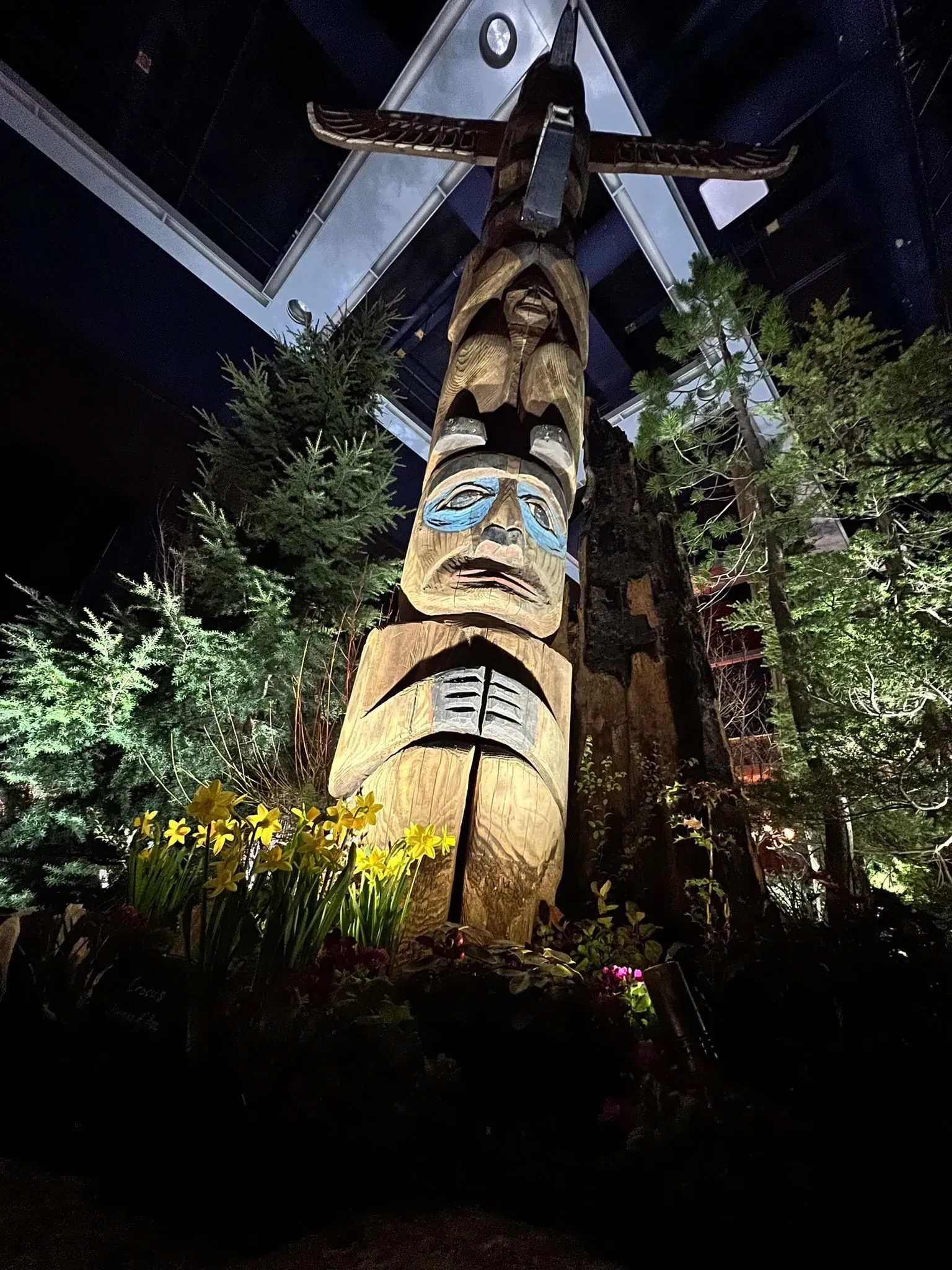 Totem pole with carved figures, lit by spotlights, surrounded by trees and daffodils.