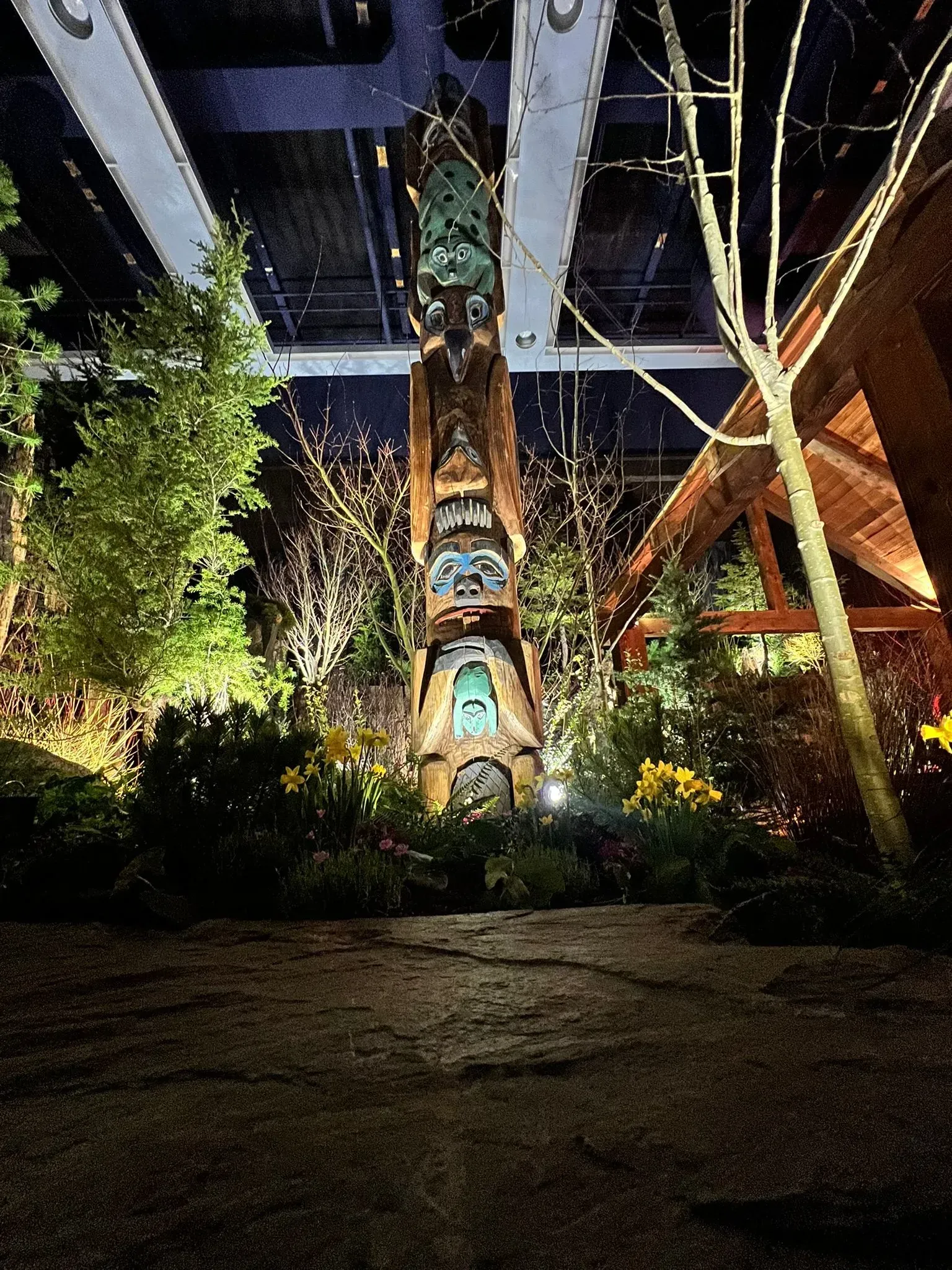 Totem pole lit at night, surrounded by plants and a building.