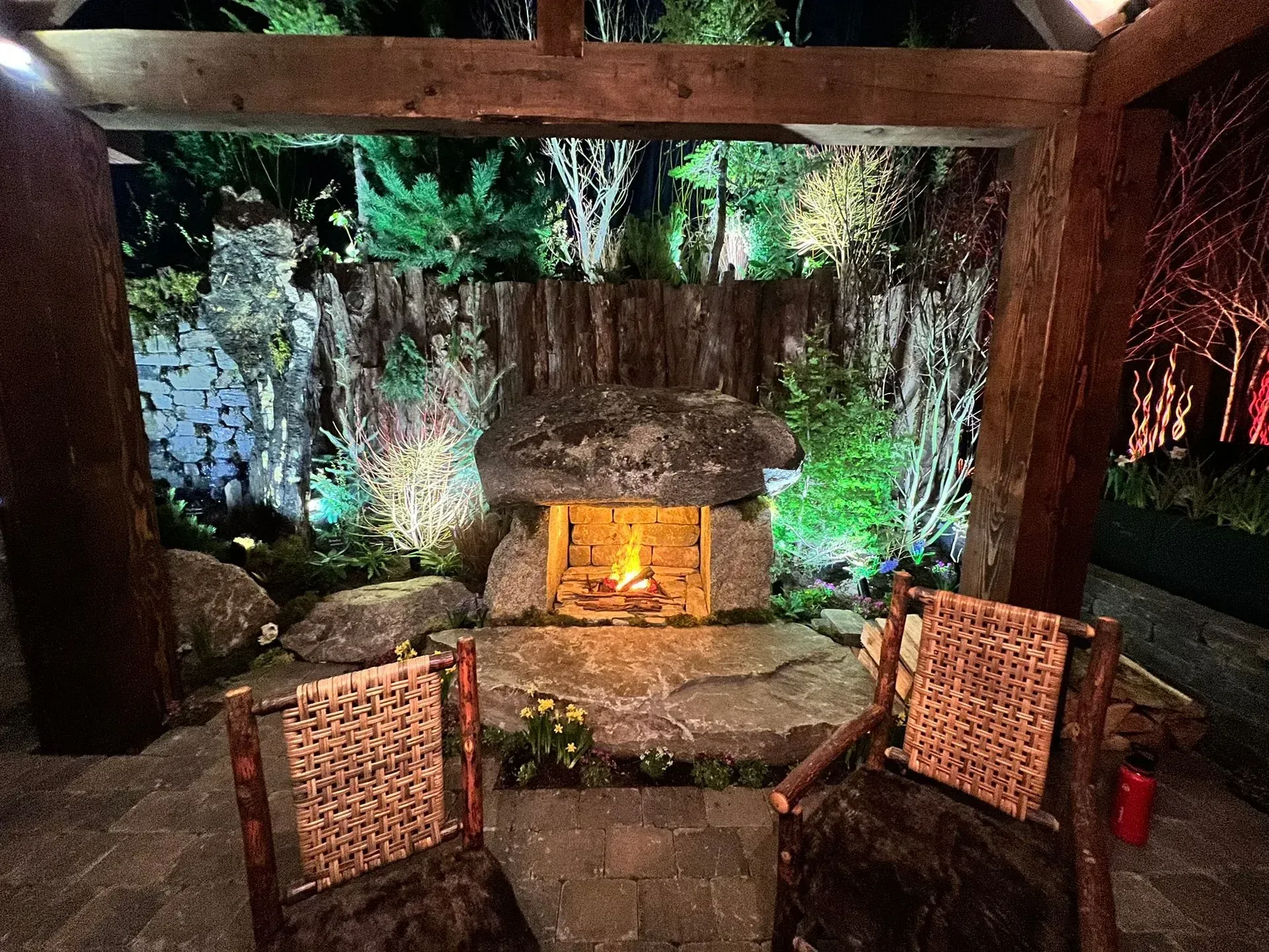 Outdoor stone fireplace with two wicker chairs. Wood beams and background greenery lit by warm light.