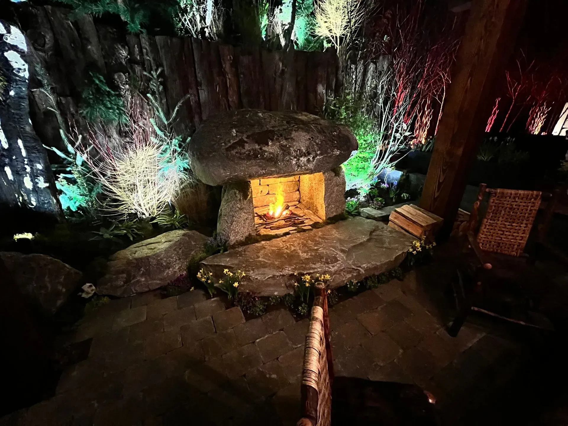 Stone fireplace with lit fire, set in a forest, surrounded by illuminated trees, with seating nearby.