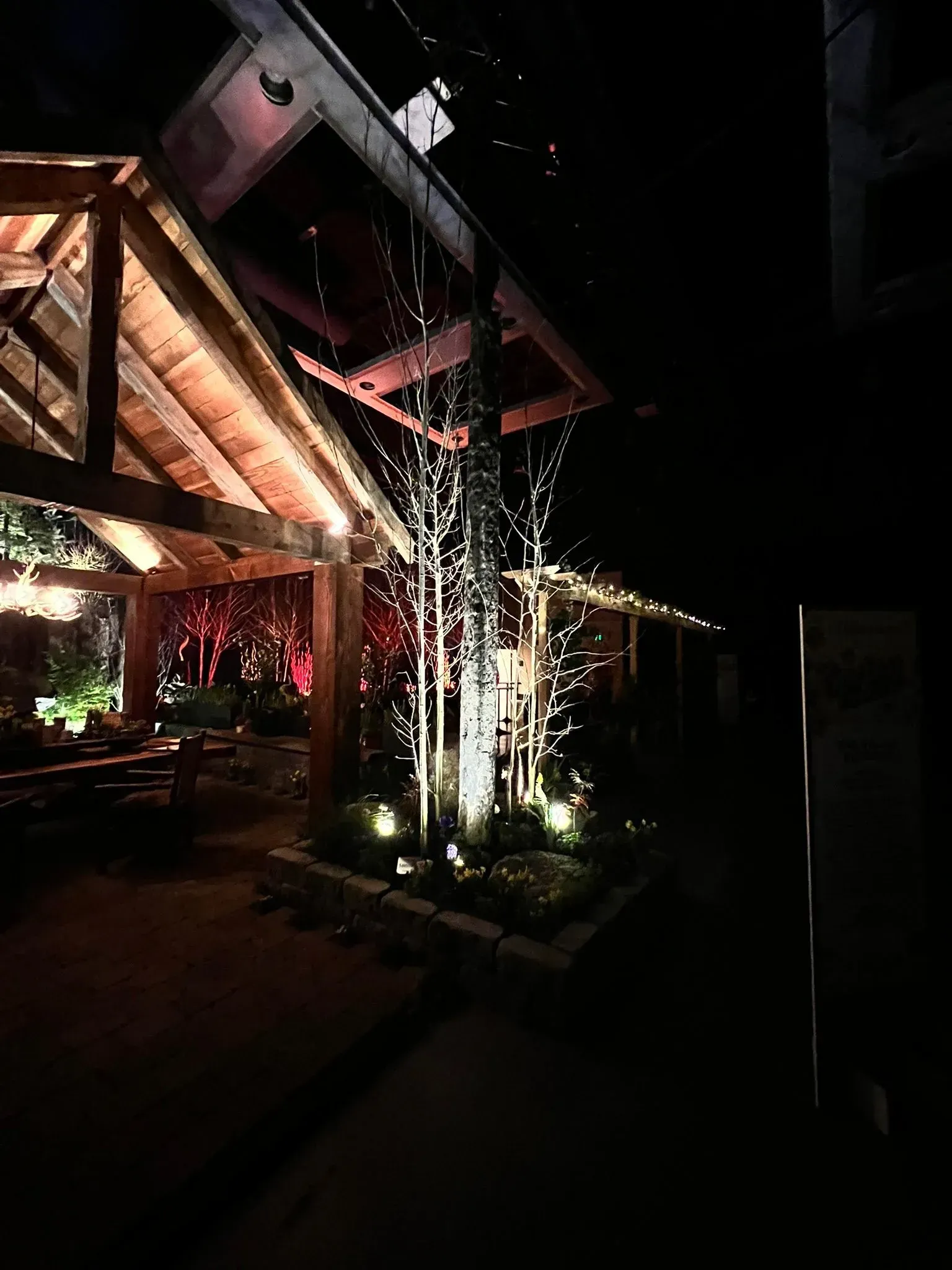 Nighttime outdoor restaurant patio with illuminated wooden beams, water fountain, and low-level garden lights.