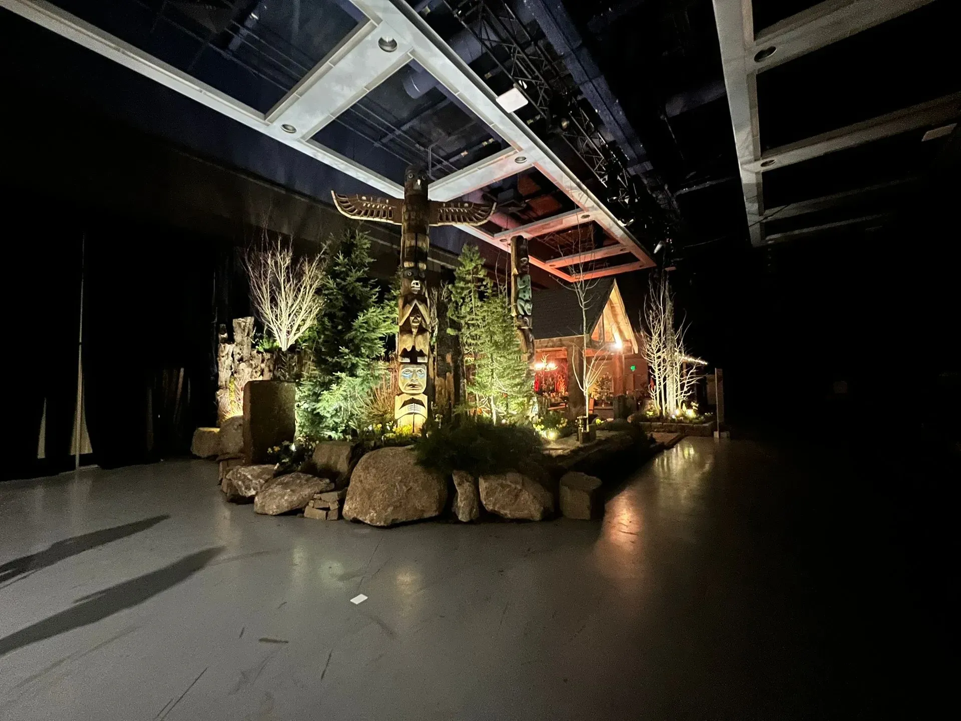 Stage set with totem pole and cabin surrounded by trees and rocks, illuminated by stage lighting.