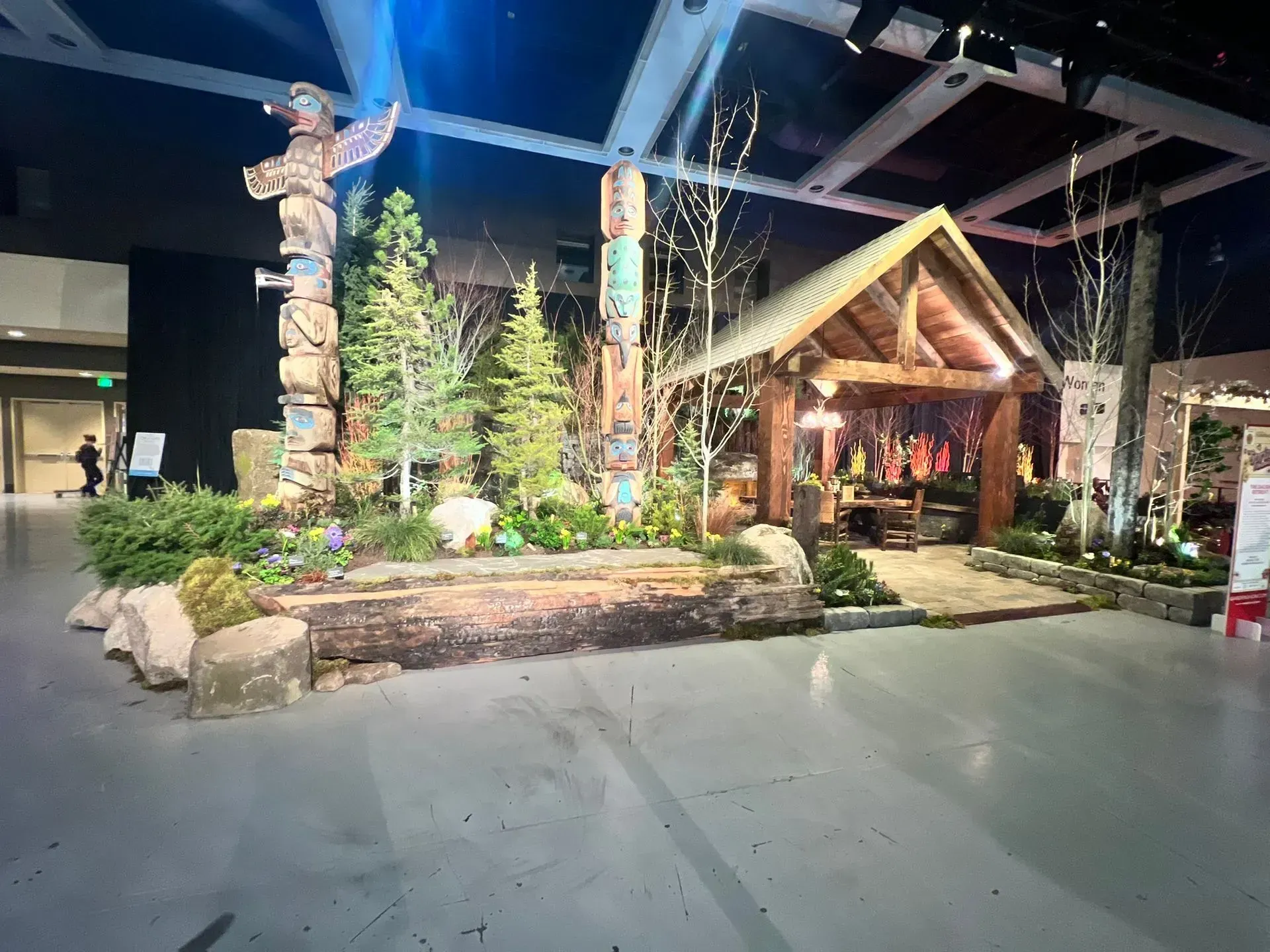 Display of totem poles and cabin scene at an event with plants and trees.