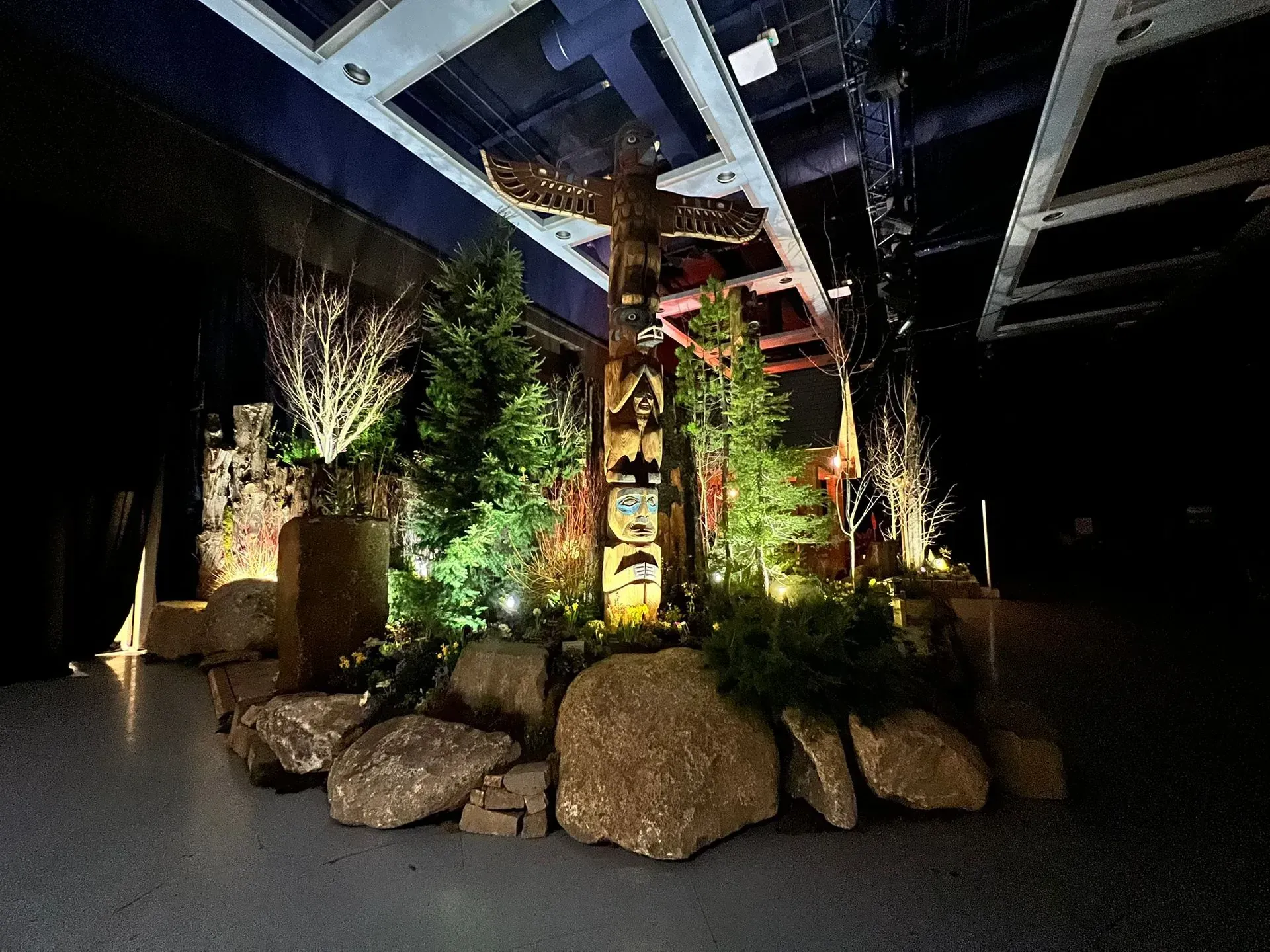 A totem pole surrounded by greenery and rocks, illuminated indoors.