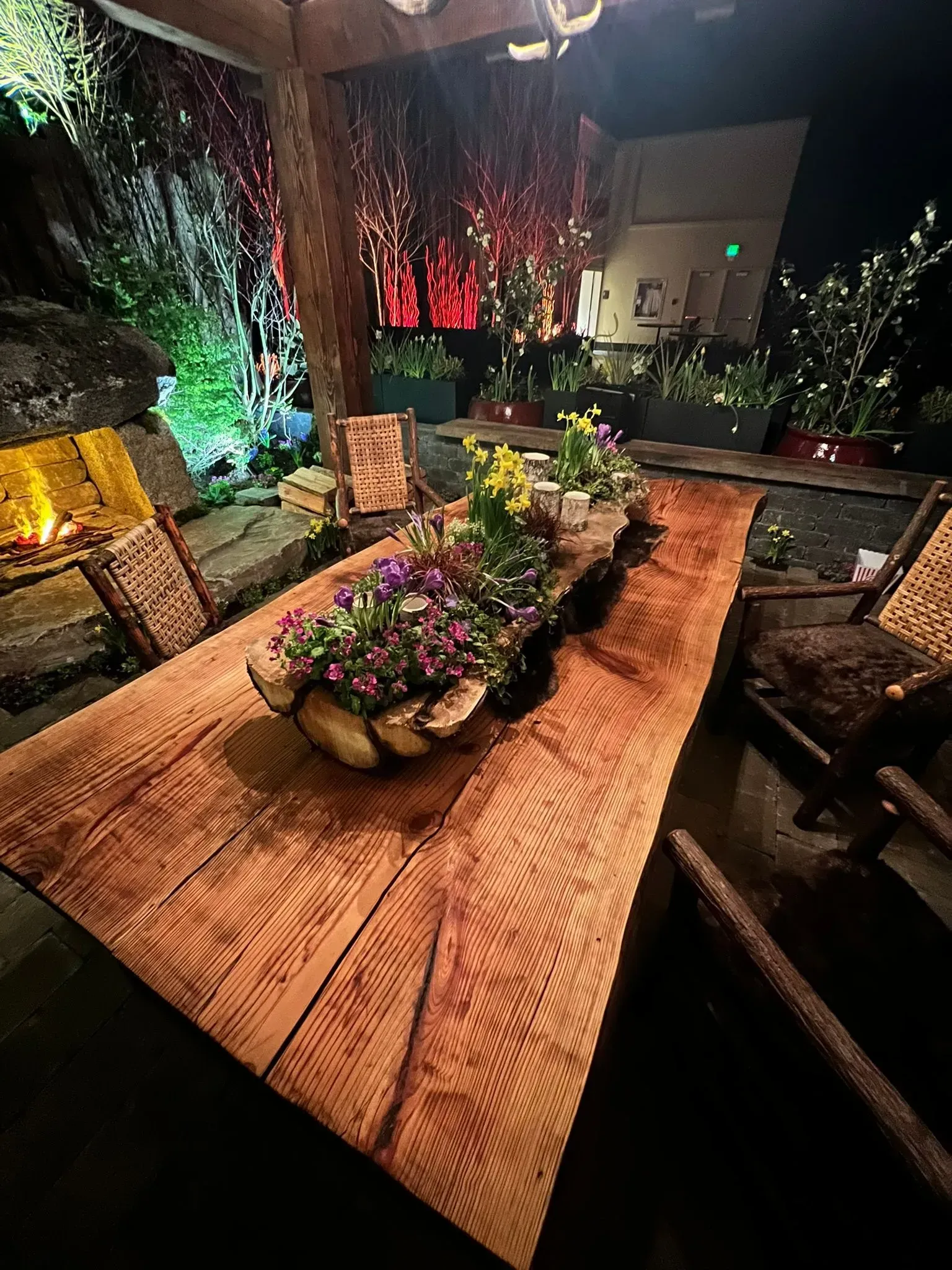 Rustic outdoor dining table with floral centerpiece, surrounded by chairs and greenery. Nighttime setting.