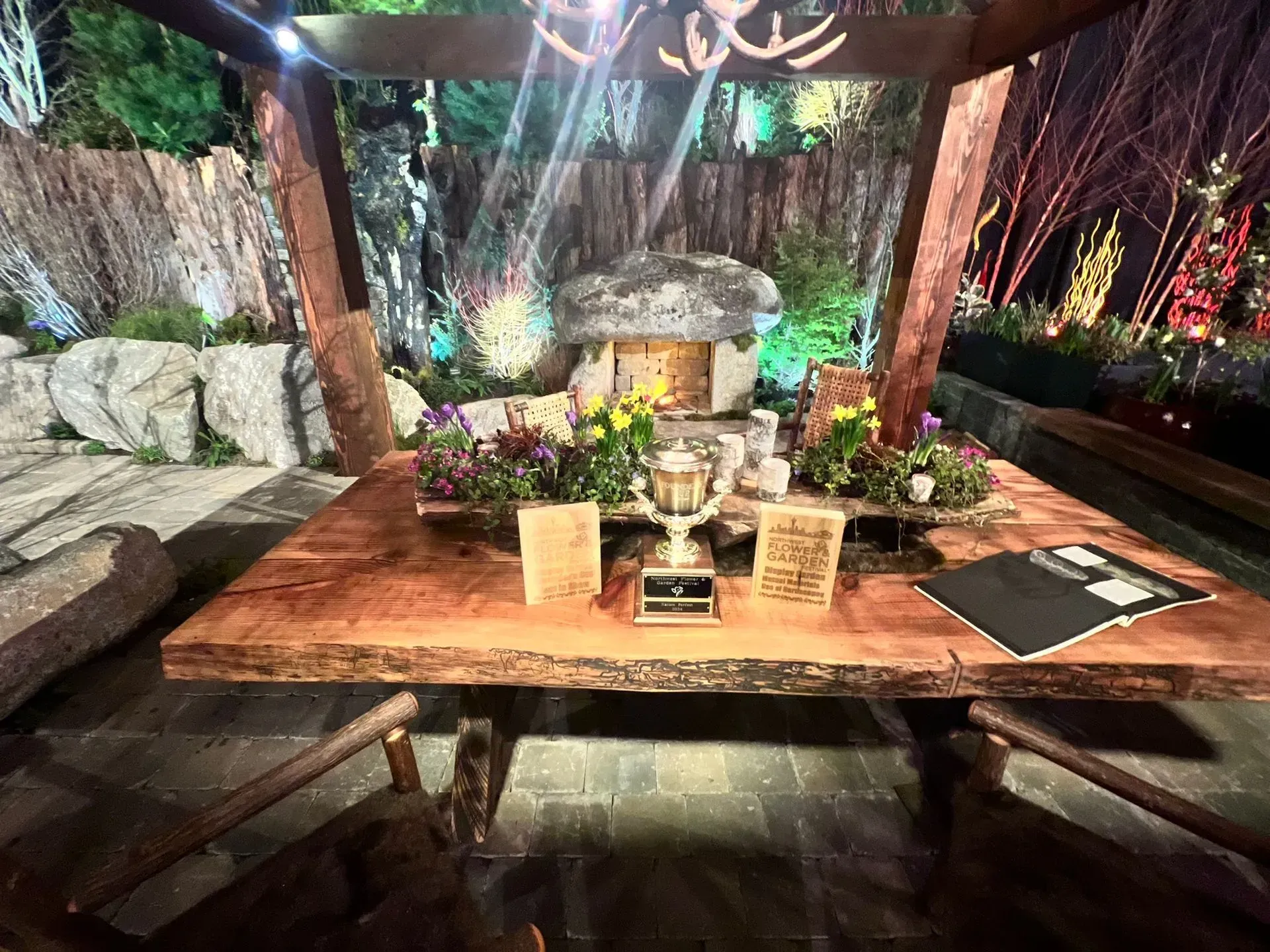 Wooden table set for display with flowers and a trophy under an illuminated gazebo in a garden setting.