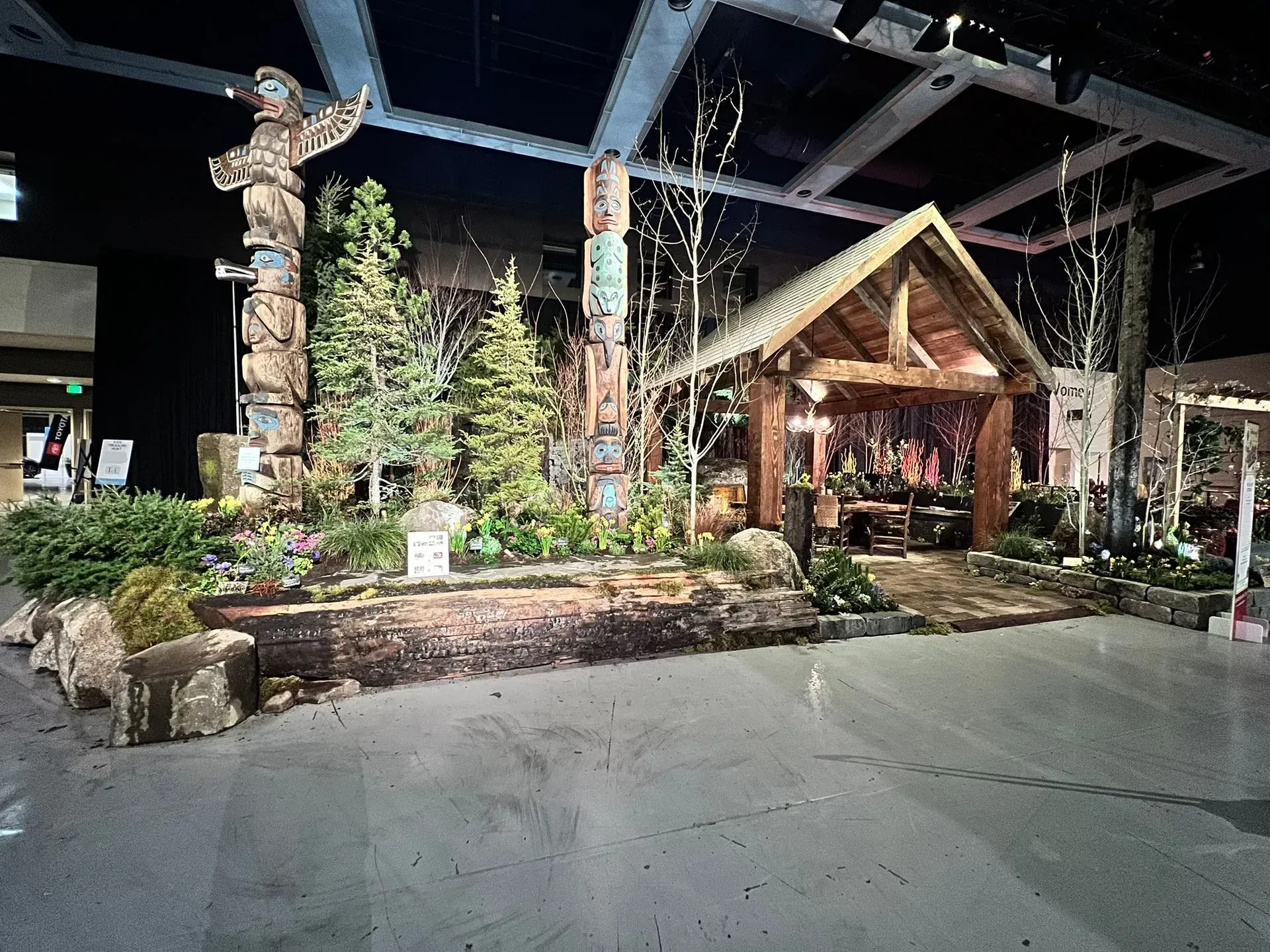 Display featuring totem poles, a cabin, trees, and plants in a large, open space.
