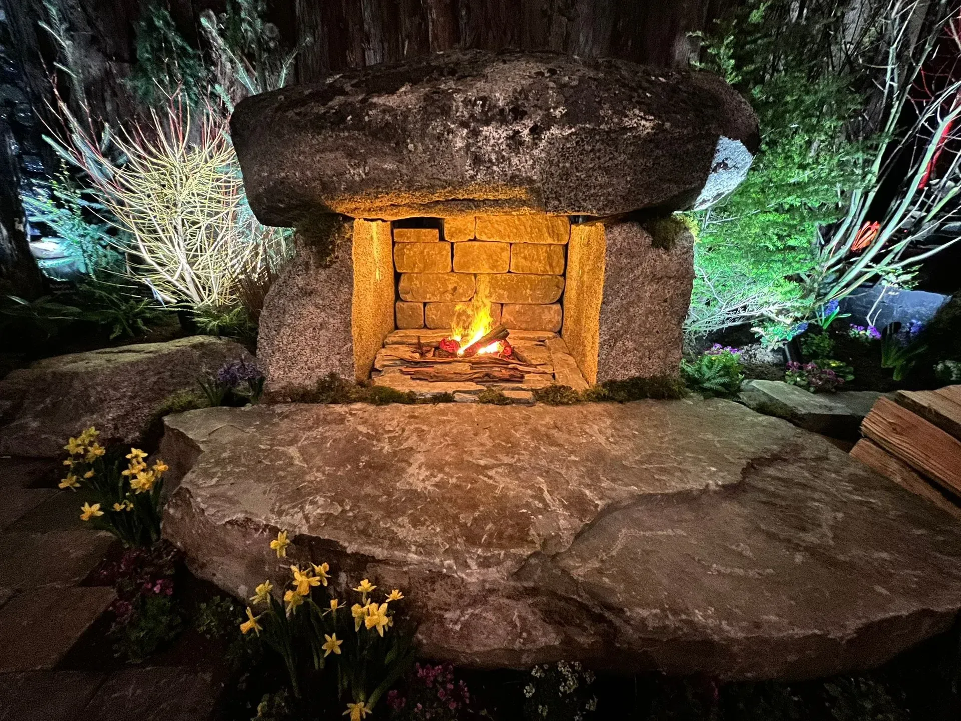 A stone fireplace with a fire, surrounded by rocks, grass, and yellow flowers, lit at night.