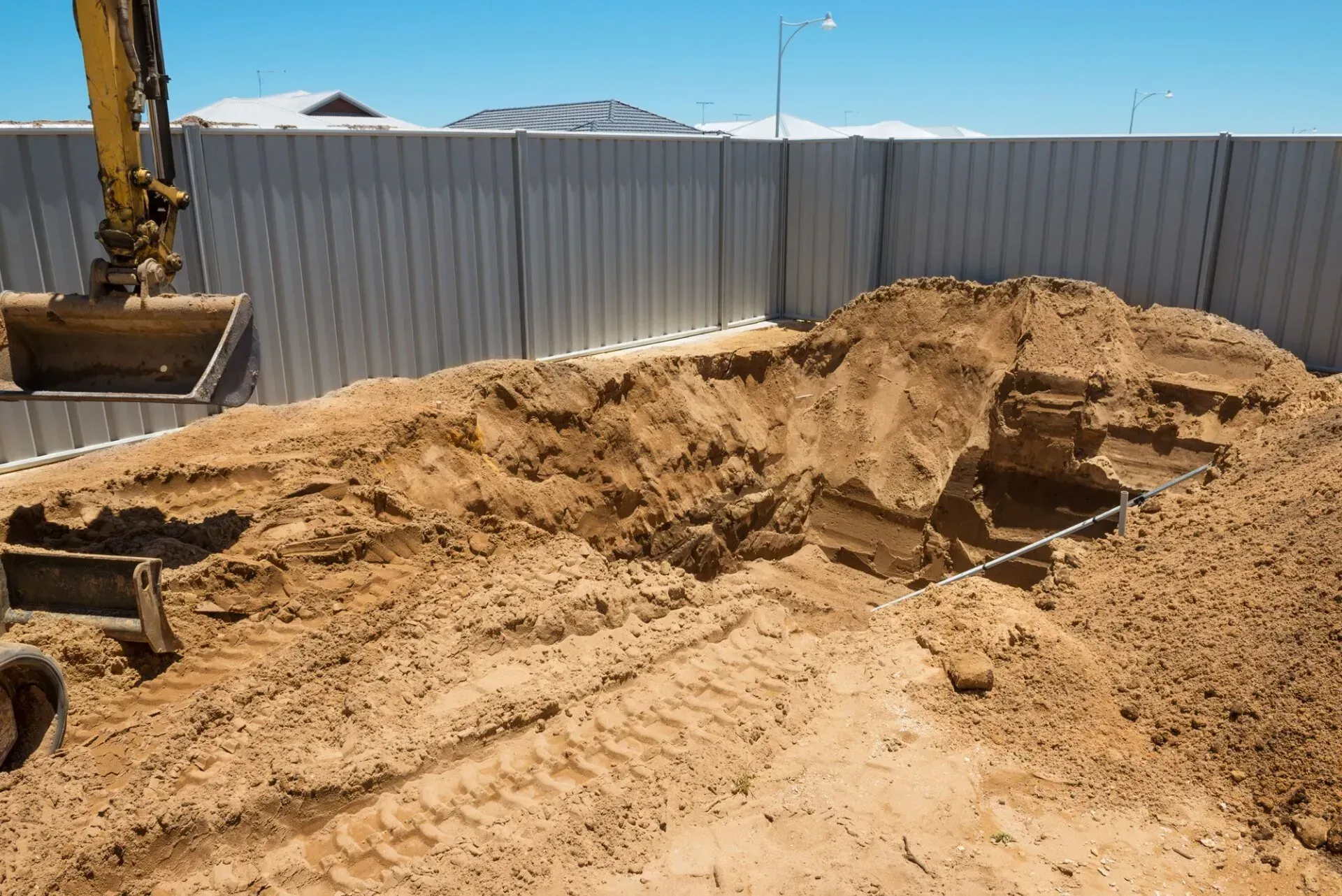 Excavator digging a trench in sand next to a grey fence under a clear sky.