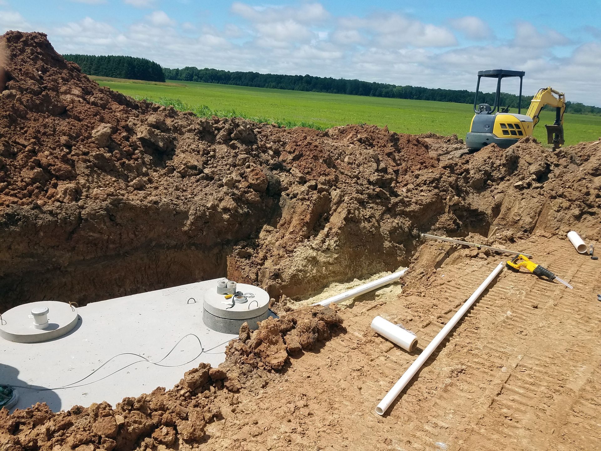 Excavation site with a septic tank; a small excavator and pipes are visible. Brown soil and green field in the background.