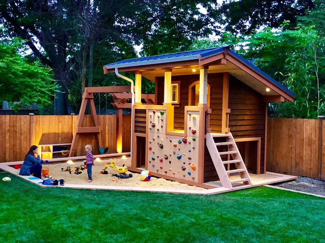 Playhouse with climbing wall, swing set, and sandpit in a backyard. Two children play.