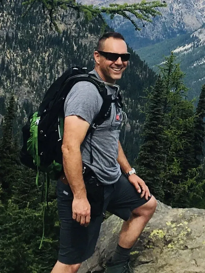 Man with sunglasses and backpack hikes on a mountain path, smiling. Green trees, and a mountain range in background.