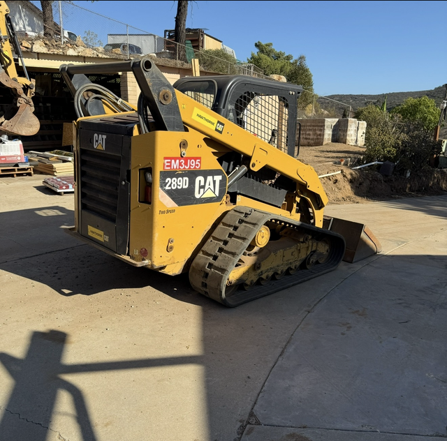 Yellow CAT 2890 tracked skid-steer on a concrete driveway near a truck and stone wall