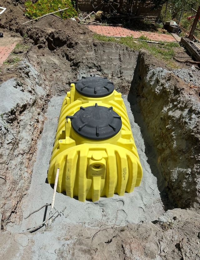 Yellow underground tank with two black lids, set in a narrow excavation trench outdoors.