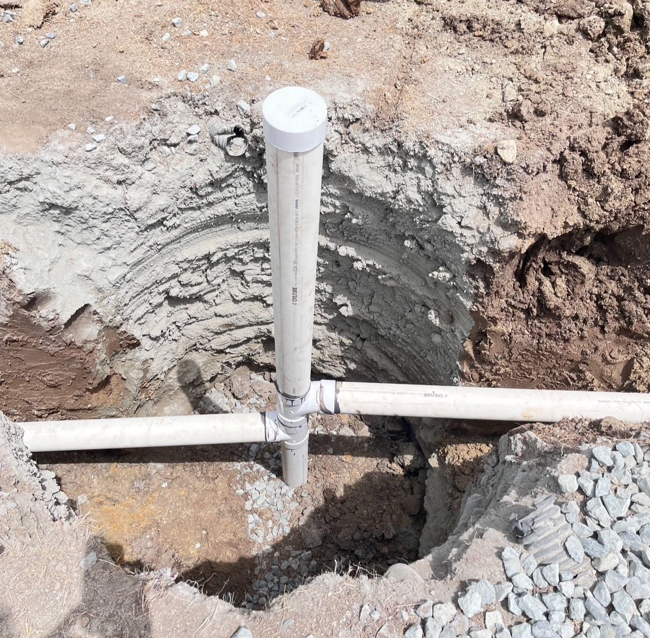 White PVC pipe junction in a dirt trench with a vertical vent pipe and two horizontal pipes.