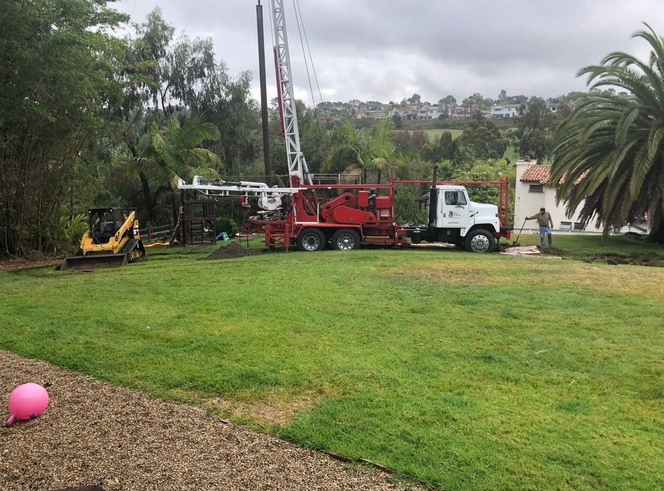 Red drilling truck and white truck parked on a grassy field near trees and a house