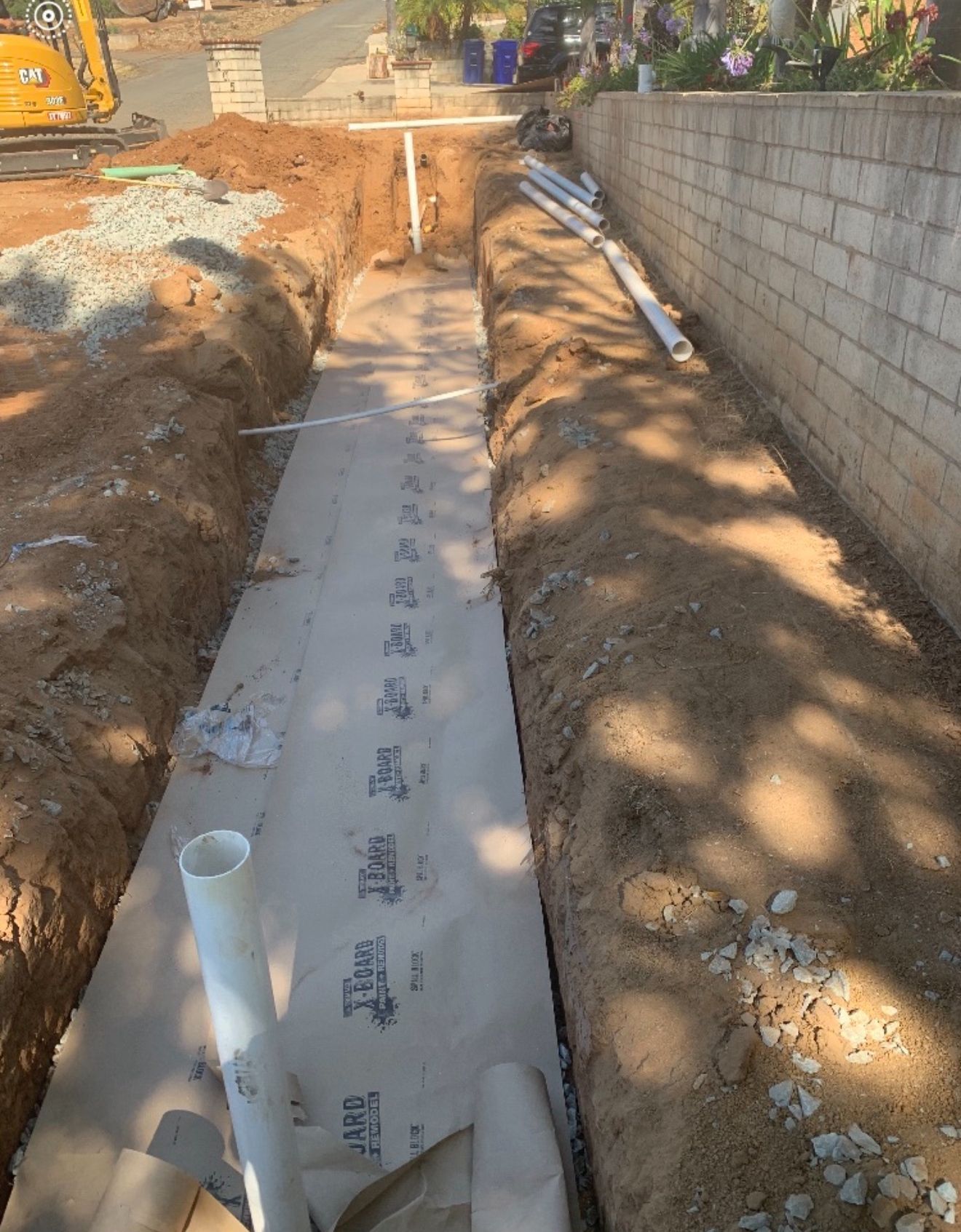 Narrow concrete walkway under construction beside a cinderblock wall, with exposed dirt and PVC pipes.