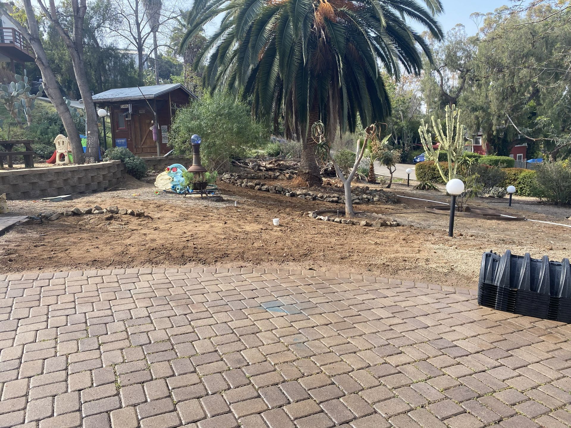 Brick patio beside a garden with palm trees, a dirt patch, and a small shaded structure.