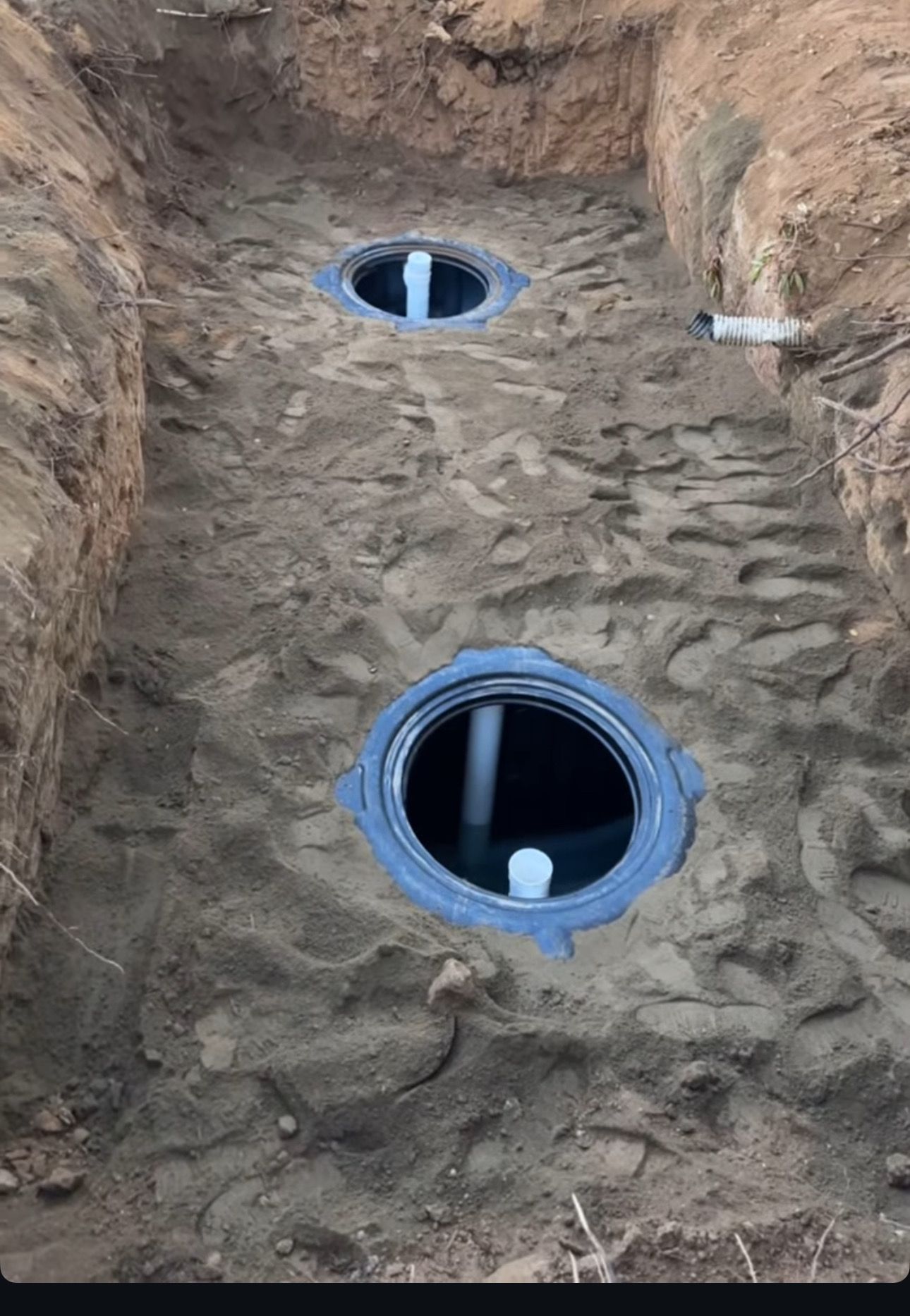 Two circular pipe openings set into a dirt trench wall, one larger with a white cap inside, one smaller above.