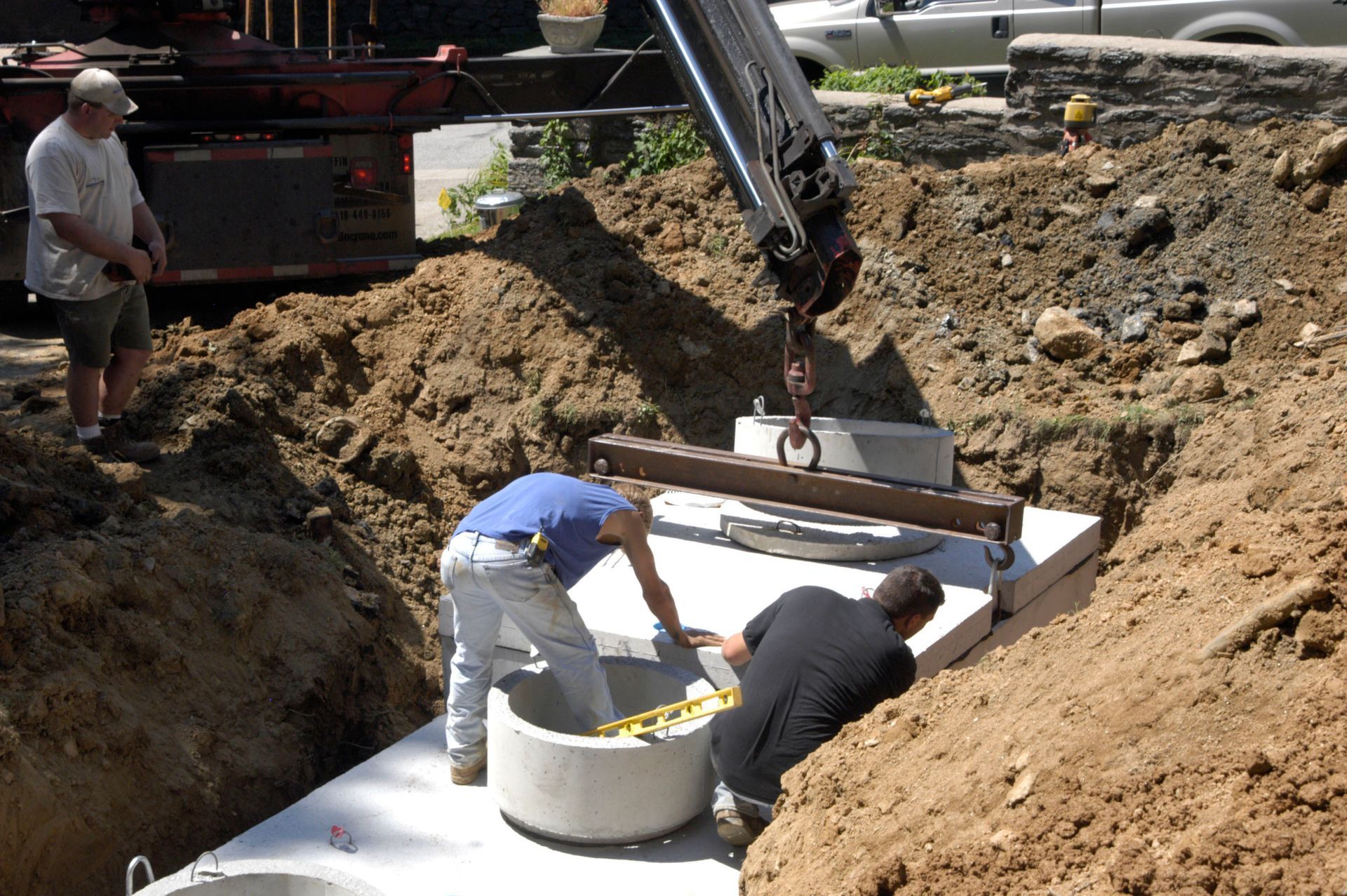 Workers installing a white septic tank in a dirt excavation with a backhoe nearby
