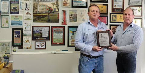 Two men hold a framed plaque in a wall of certificates and awards.