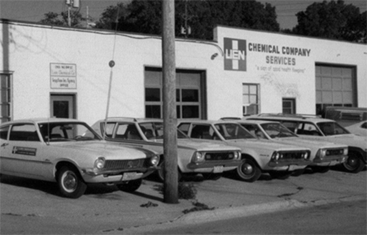 Black-and-white street scene with four parked vintage cars outside a chemical company building.