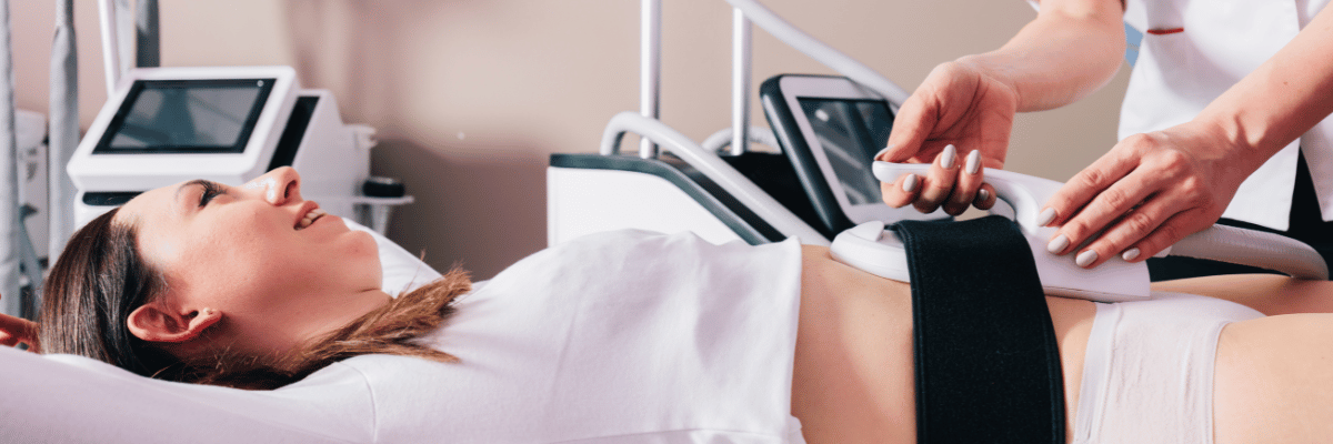 A woman receiving a body contouring treatment. A professional applies a device to her abdomen while she reclines on a table.