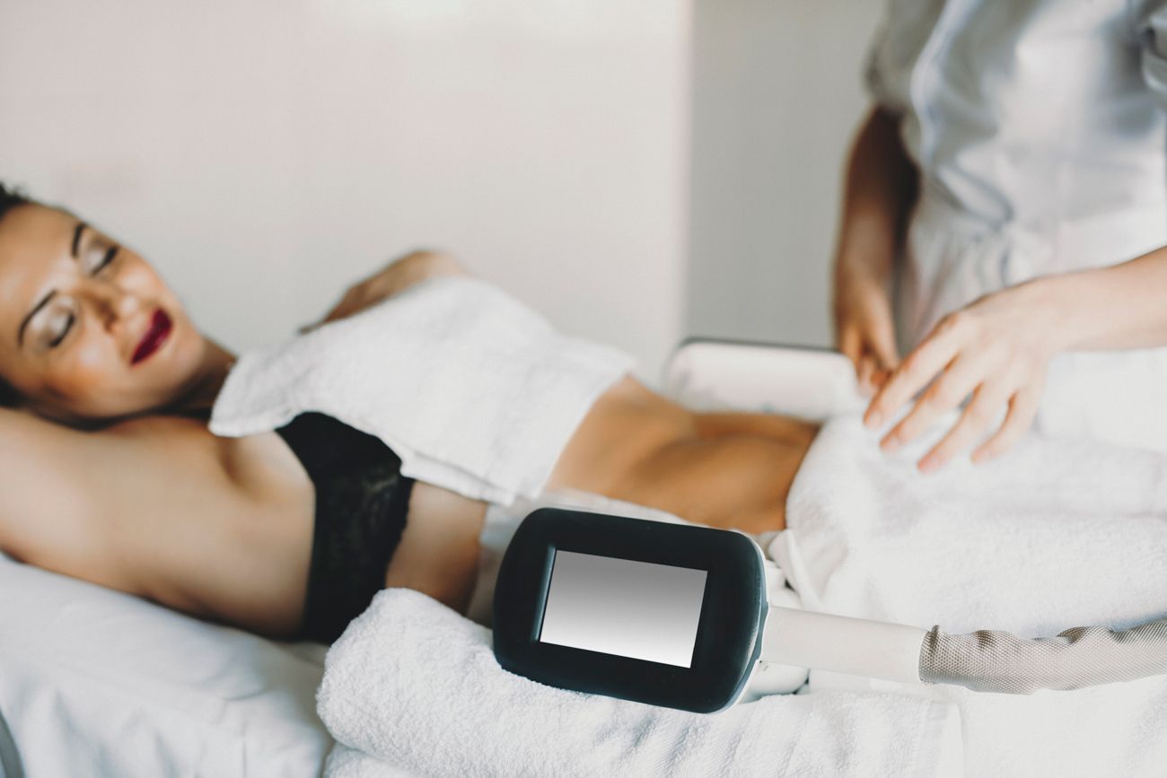 Woman undergoing a body contouring procedure; lying on a bed with a technician, device on abdomen.