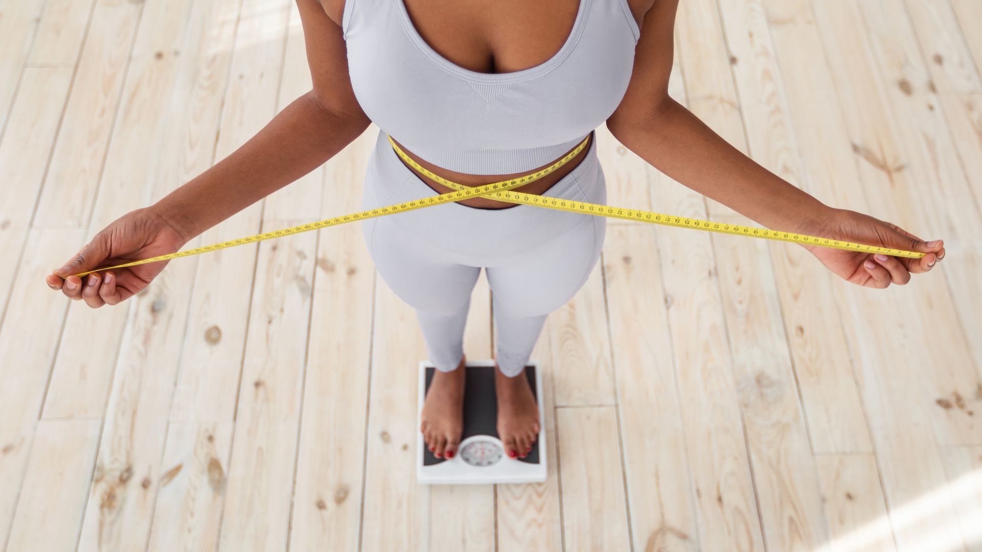 A woman stands on a scale, measuring her waist with a yellow tape. She wears a light grey athletic outfit on wooden flooring.