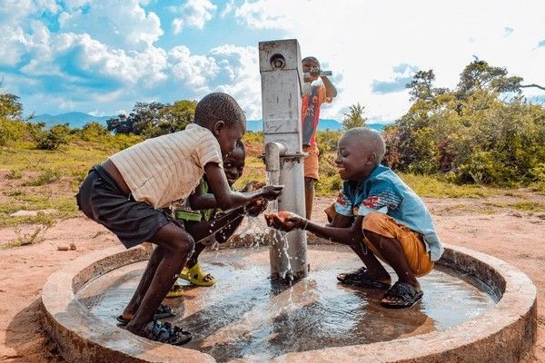 Children washing hands at a water pump outdoors. One boy smiles, two others look focused. Sunny day.