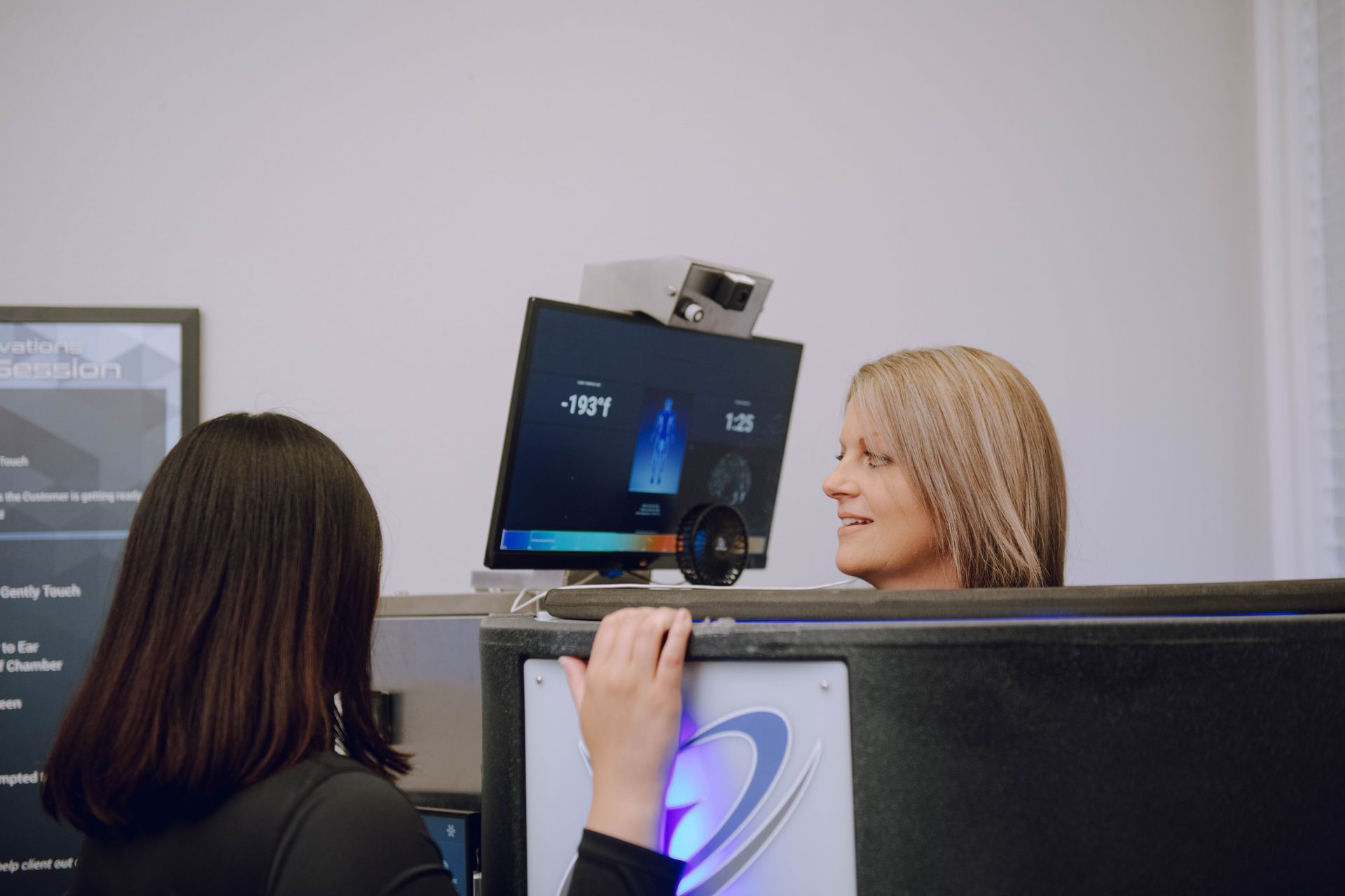 Woman inside a cryotherapy machine, talking to an attendant at a medical clinic. The screen displays data.