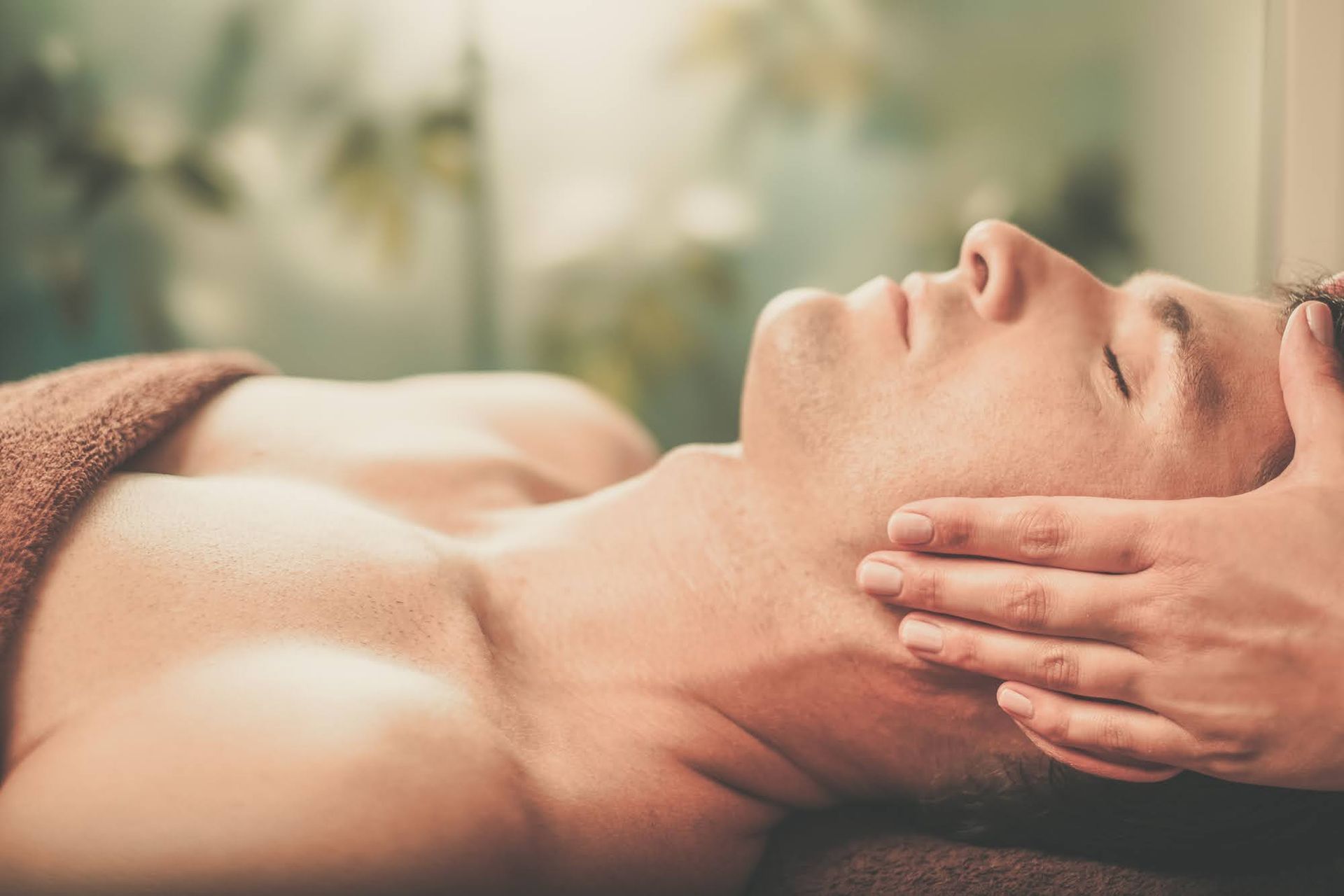 Man receiving a massage, face up with eyes closed, hands on face and neck. Brown towel, blurred background.
