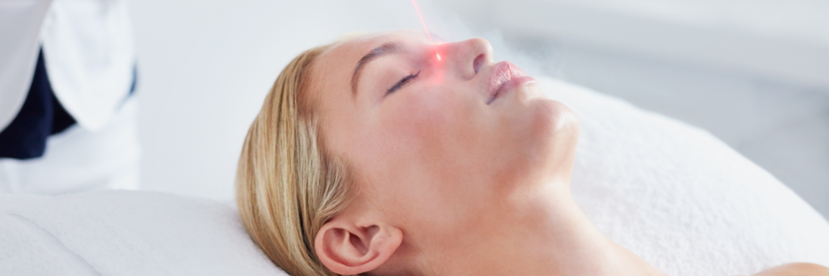 Woman receiving laser treatment on her nose, lying down with eyes closed. Red laser light visible.