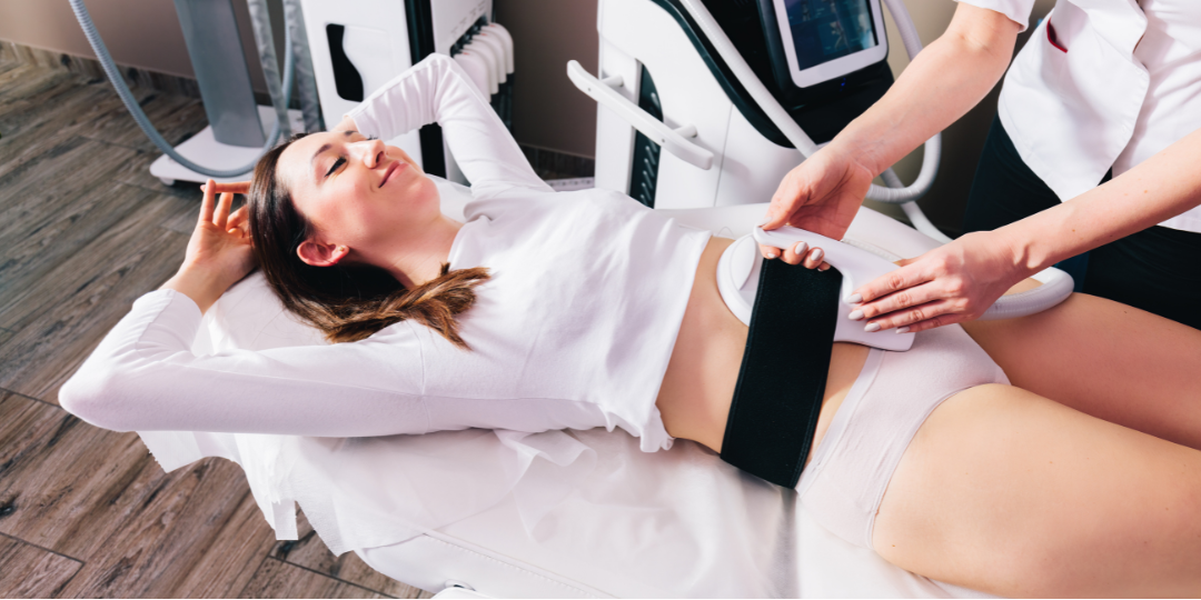 A woman lies on a table, receiving a body sculpting treatment. A technician holds a device to her abdomen.