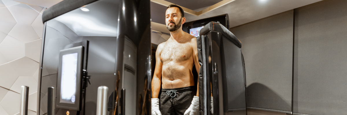 A shirtless man wearing gloves stands in front of a body scanner in a modern room. He looks upward with a neutral expression.