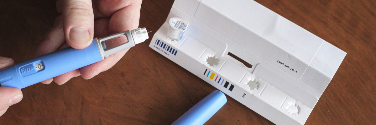 A person's hands holding a blue insulin pen, with the cap partially removed, next to a white tray containing insulin cartridges.