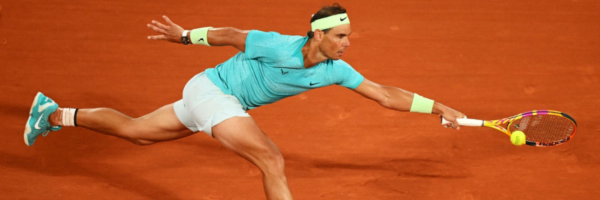Rafael Nadal in action on a clay court, diving to hit a forehand. He wears a blue shirt, white shorts, and a yellow headband.