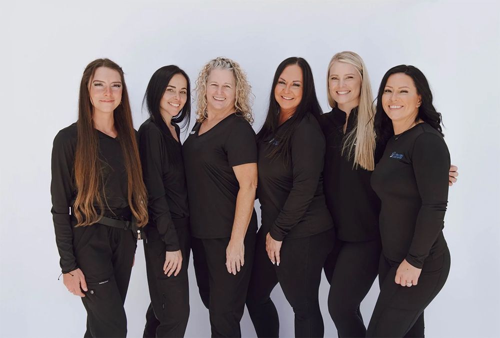 Six women in black scrubs pose against a white background, smiling at the camera.