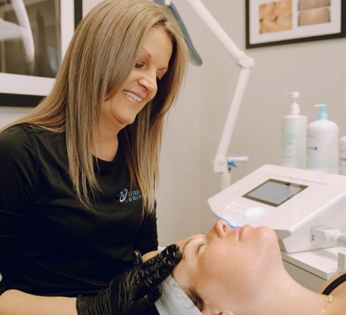 A woman in a black shirt with a logo performs a facial treatment on a client. The setting is a brightly lit spa room.